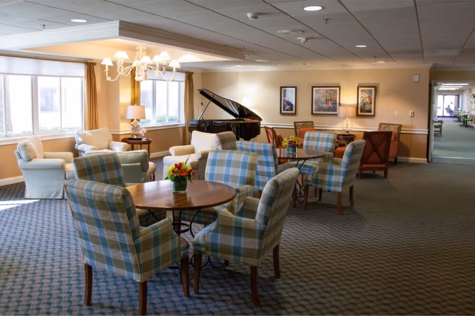 A spacious and well-lit common area with multiple round wooden tables surrounded by plaid upholstered chairs. There are comfortable armchairs and sofas near large windows with beige curtains. A black grand piano is positioned against the wall, which is decorated with framed artwork. The carpet is patterned, and the ceiling has recessed lighting and a chandelier.