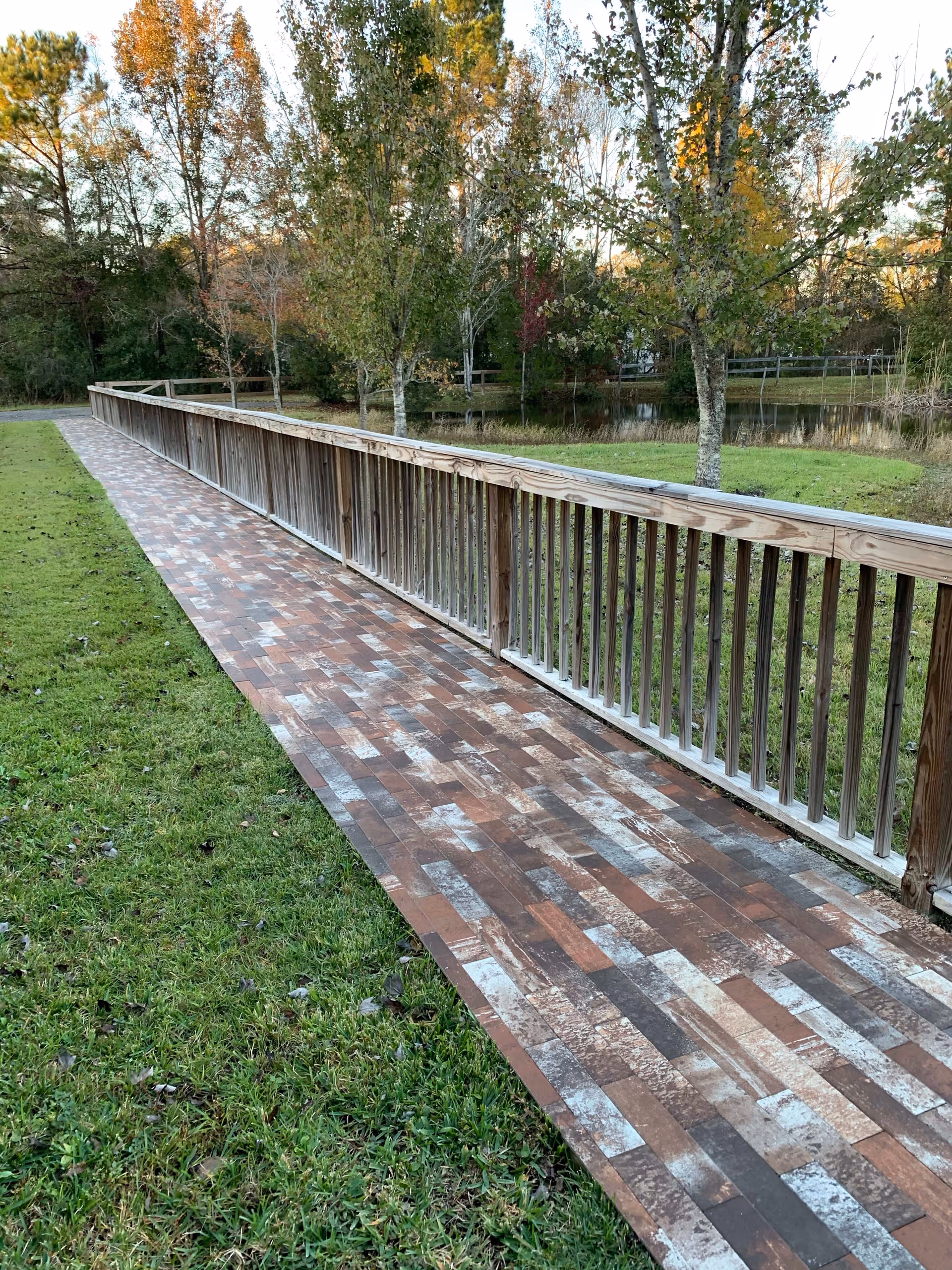 A long outdoor brick-patterned walkway with a wooden railing runs beside grass, trees, and a small pond.