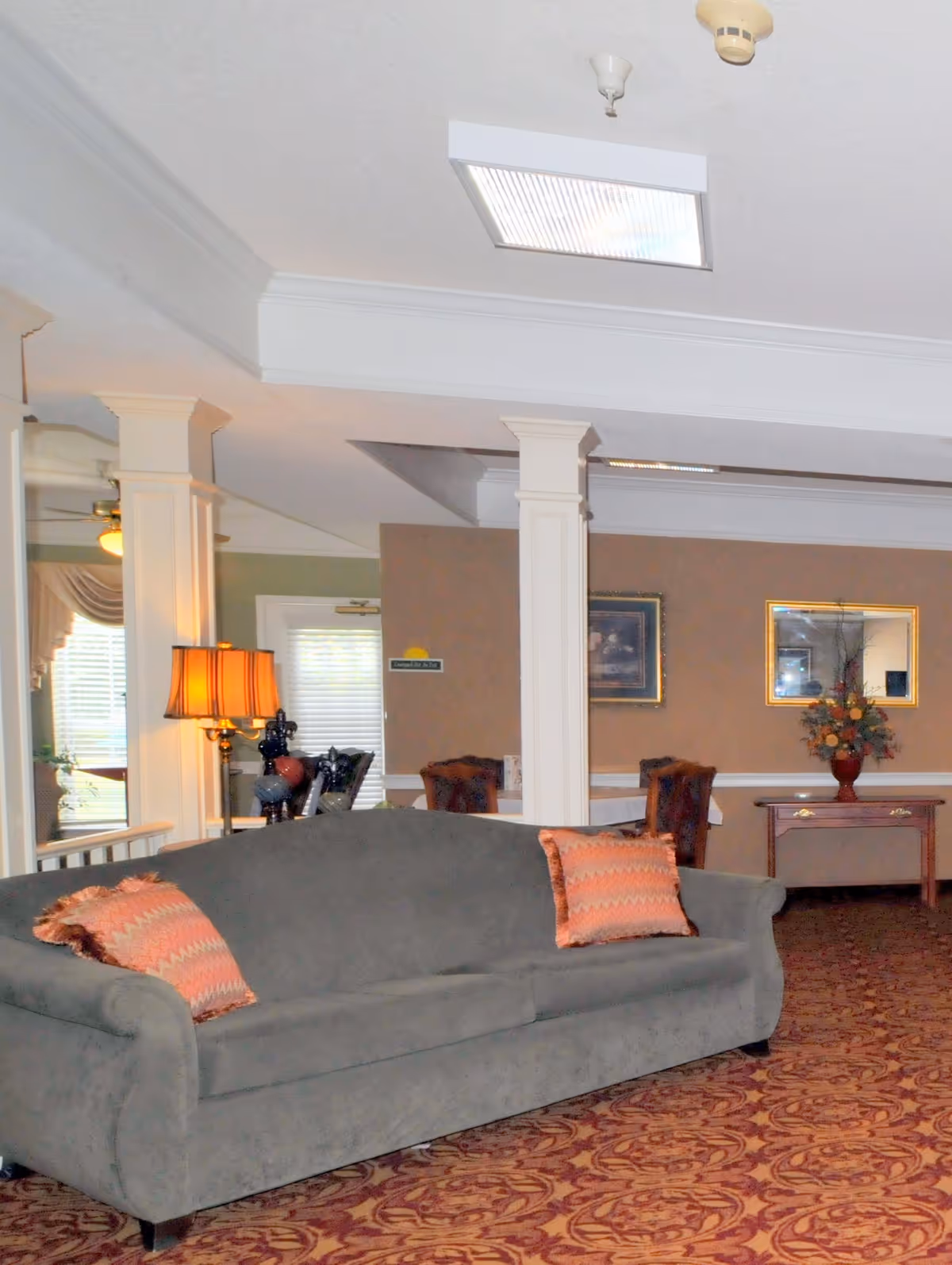 Carpeted common lounge with a gray sofa and patterned pillows, decorative columns, lamp and chairs in the background.