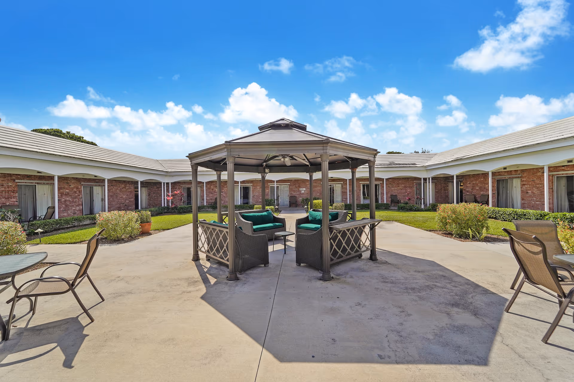 Outdoor courtyard area at Manor Pines featuring a central gazebo with green cushioned seating, surrounded by a paved walkway, patio chairs and tables, and a single-story brick building with multiple doors and windows under a blue sky with scattered clouds.