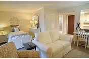 A bright and cozy living space in a senior living facility featuring a white loveseat, an armchair, a bed with decorative pillows and a headboard, a small desk with a chair, and soft lighting from table lamps. The room has light-colored walls and flooring, creating a warm and inviting atmosphere.