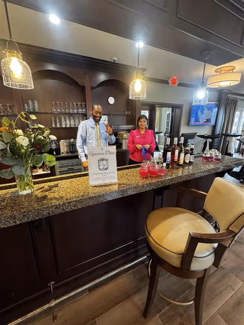 Two staff members standing behind a granite countertop bar in a senior living facility. The man on the left is smiling and making a peace sign, while the woman on the right is standing with her hands clasped. The bar has several bottles of wine and glasses filled with a pink beverage. There is a sign on the counter advertising a signature drink called Cambridge Blue Smash. The background shows shelves with glassware and a TV mounted on the wall.