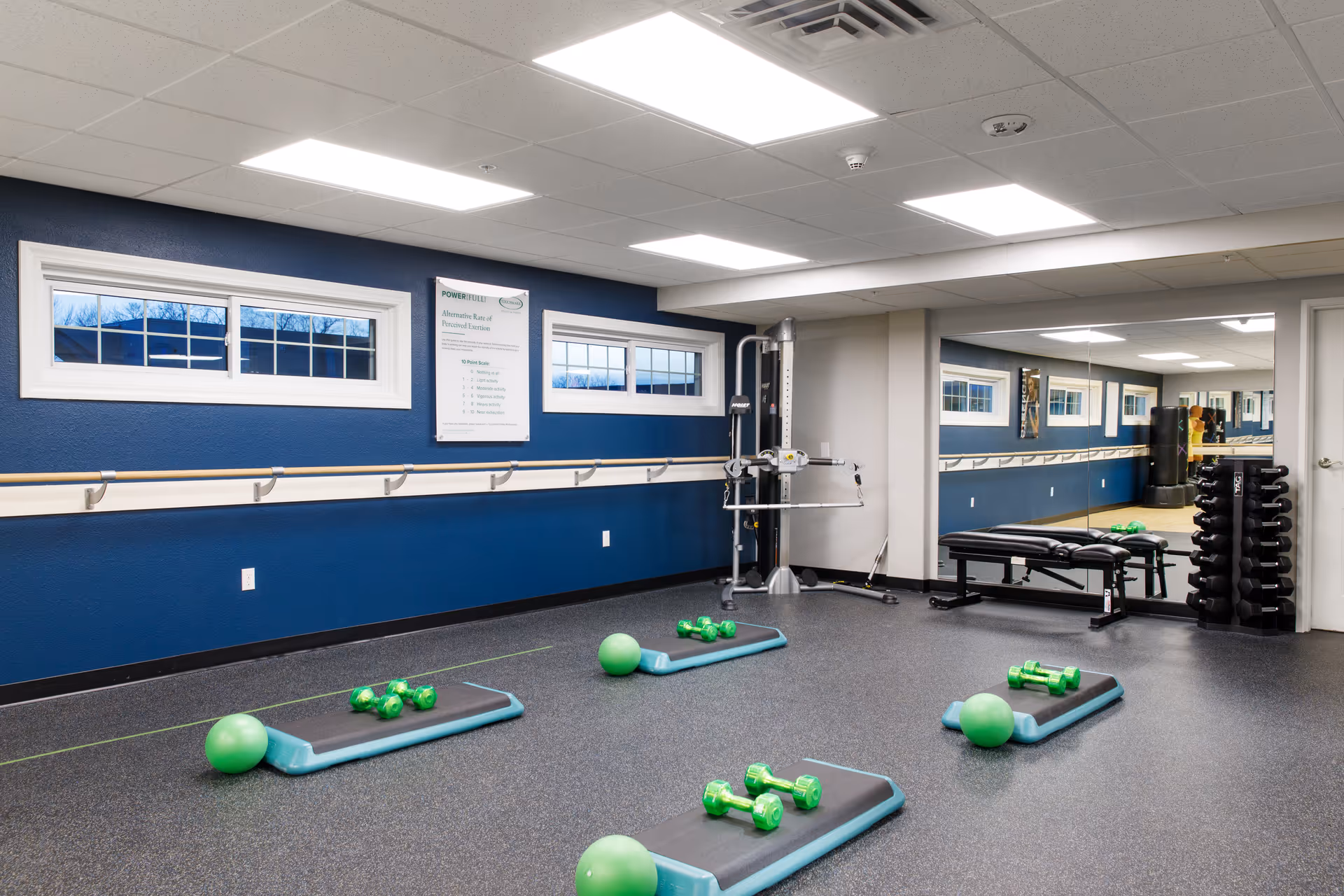 Interior fitness room with exercise mats, green dumbbells and stability balls, a mirrored wall and weight equipment.