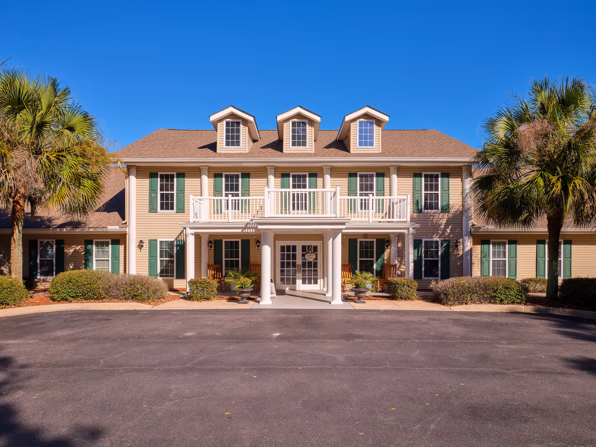 Front exterior view of a two-story building with beige siding, green shutters, and a brown roof with three dormer windows. The entrance features white columns supporting a balcony with white railings. There are palm trees and bushes on either side of the entrance, and a paved driveway in front under a clear blue sky.