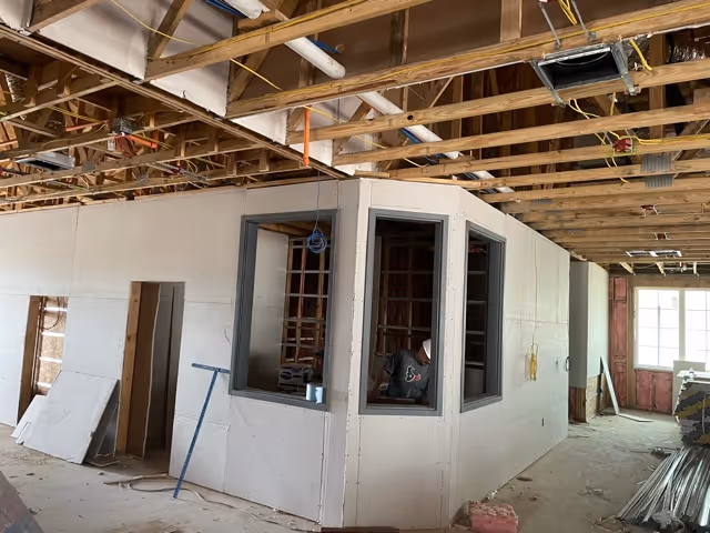 Interior view of a building under construction showing exposed wooden ceiling beams, partially finished drywall walls, and a small room with three windows. Construction materials and tools are scattered around the area.