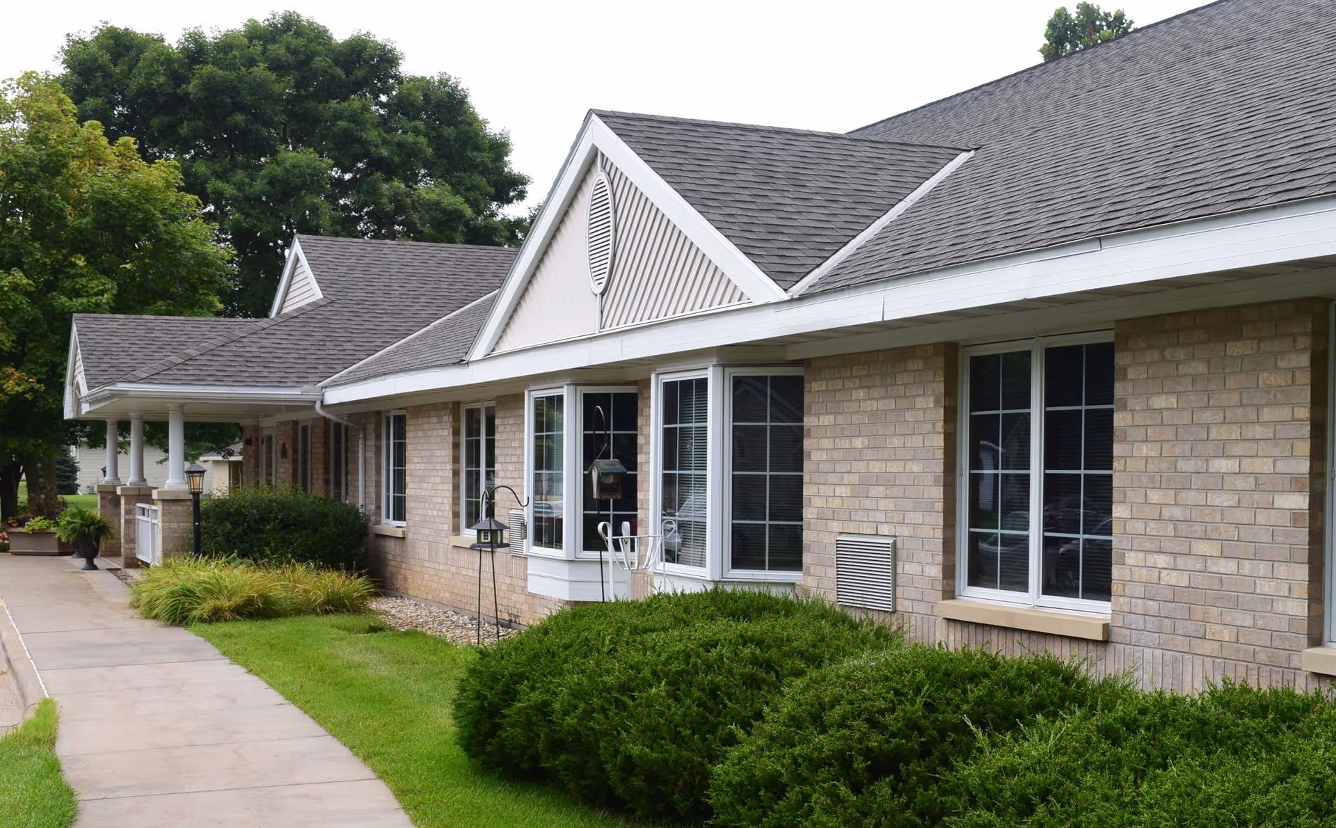 Exterior view of a single-story brick building with multiple windows, a covered porch, and well-maintained landscaping including bushes and trees along a concrete walkway.