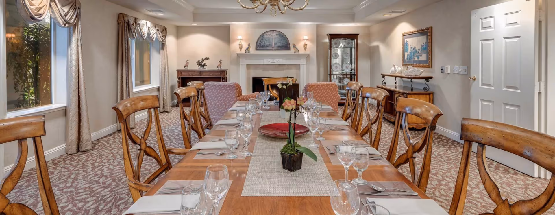 A formal dining room with a long wooden table set for a meal, including glassware, napkins, and cutlery. The room features wooden chairs, two upholstered armchairs at the far end, large windows with draped curtains, a fireplace with decorative items on the mantel, a glass display cabinet with plates, and framed artwork on the walls. The carpet has a leaf pattern and the room is softly lit with wall sconces and a chandelier.