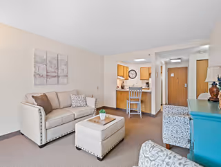 Furnished living area with a sofa, ottoman, armchairs, a small kitchen/bar, and a hallway leading to the front door.