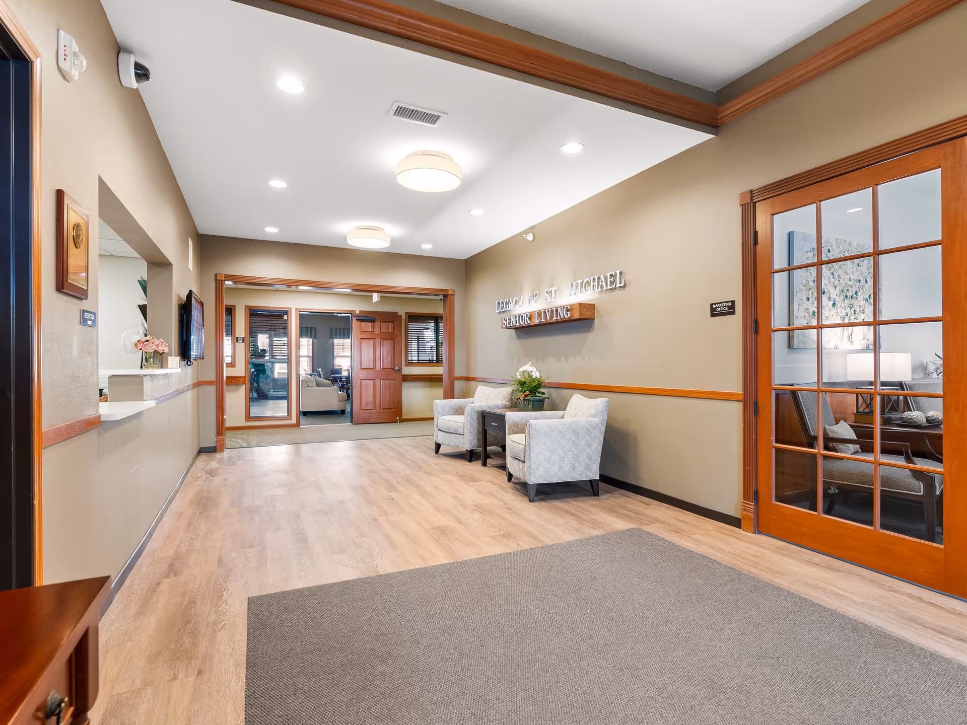 Reception lobby of a senior living facility with seating, a reception window, wood‑trimmed doors, and wall signage.