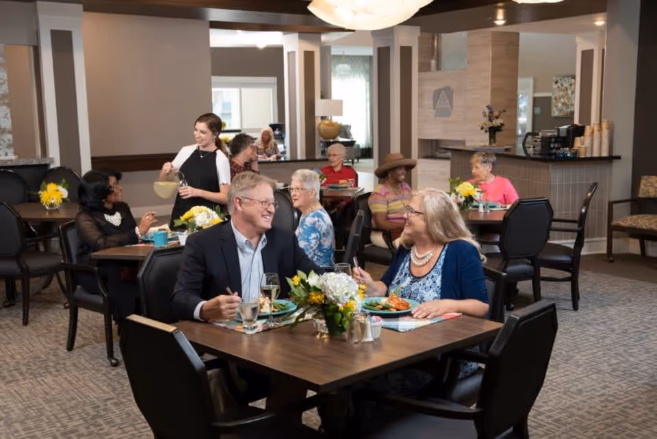 A dining room in a senior living facility with several elderly residents seated at tables enjoying meals and conversation. A staff member is pouring a drink for one of the residents. The room is warmly lit and decorated with flowers on the tables.