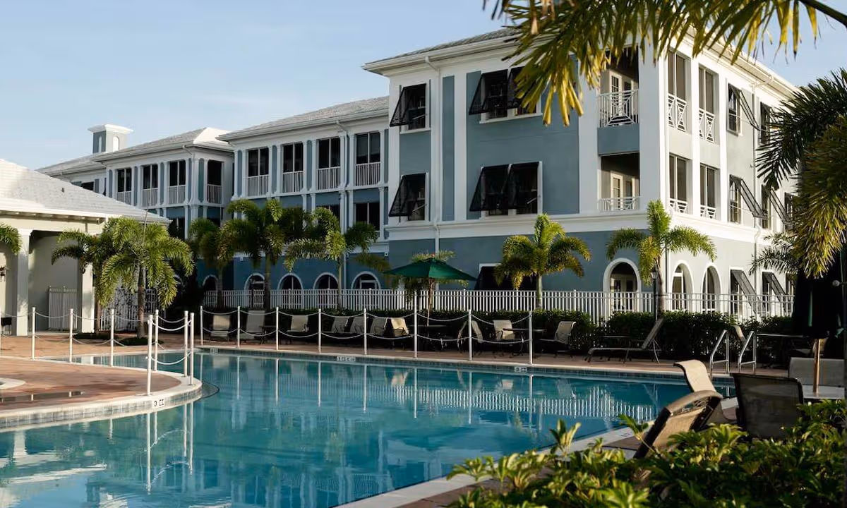 Outdoor swimming pool area with lounge chairs and palm trees in front of a multi-story residential building with balconies and black window awnings under a clear sky.