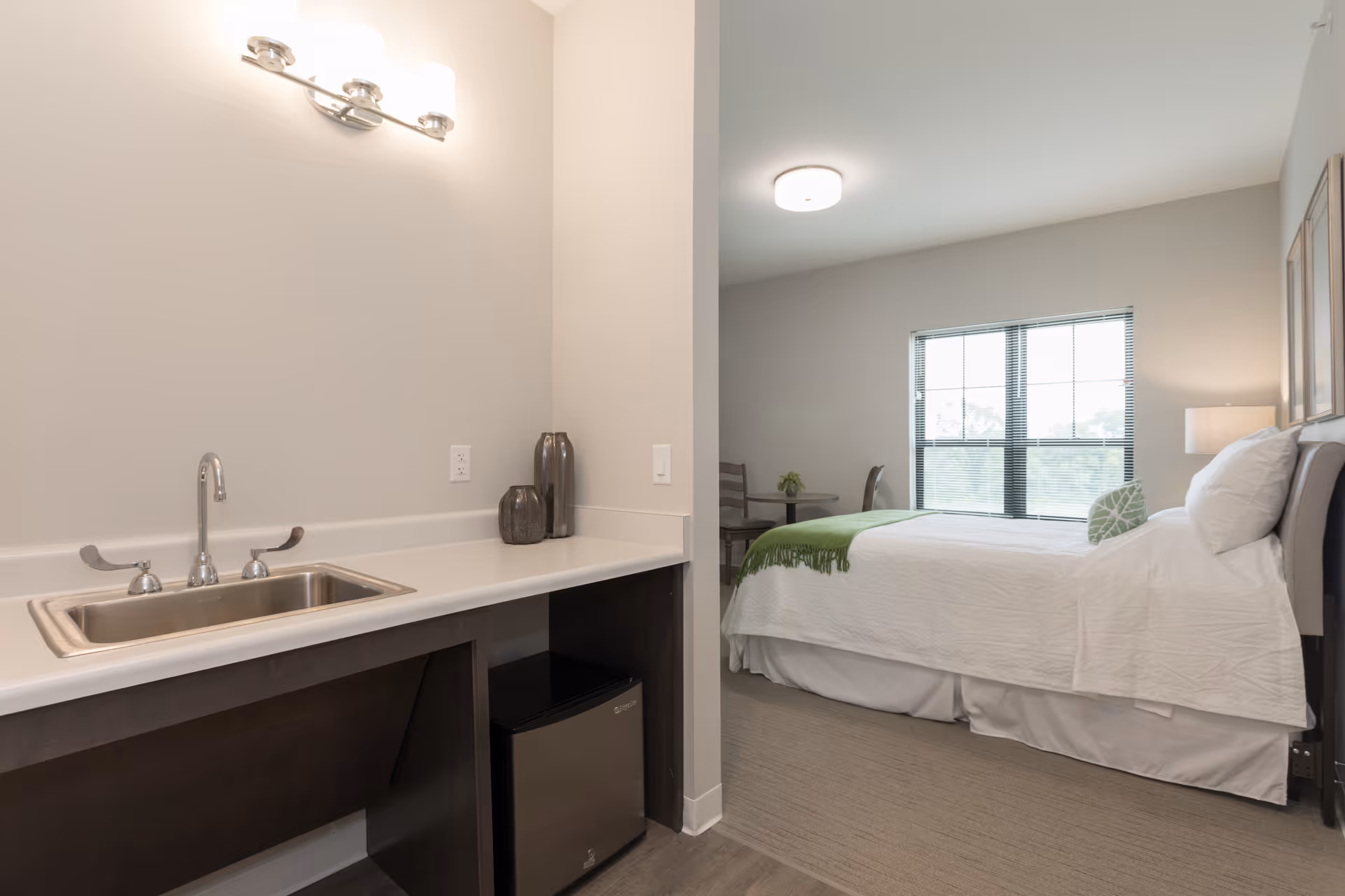 A senior living facility room featuring a small kitchenette area with a sink, countertop, and mini refrigerator. Adjacent to the kitchenette is a bedroom with a neatly made bed, white bedding, a green throw blanket, a pillow, a window with blinds, a small table with chairs, and a lamp on a nightstand.