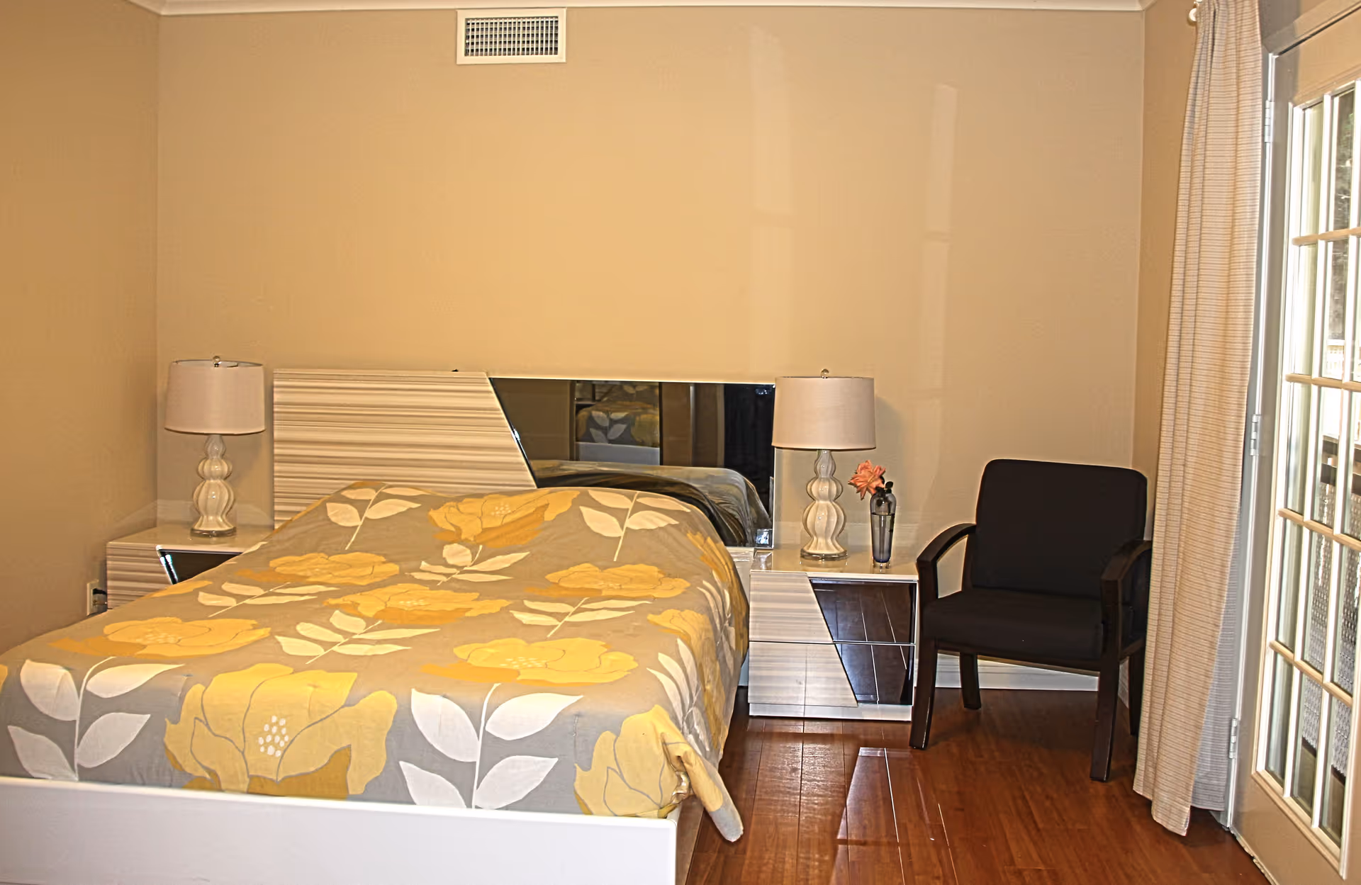 A bedroom with a bed covered in a yellow and gray floral patterned bedspread. The bed has a modern headboard with a mirror panel. On each side of the bed are matching nightstands with white lamps. To the right of the bed is a dark armchair next to a window with beige curtains and a glass door.