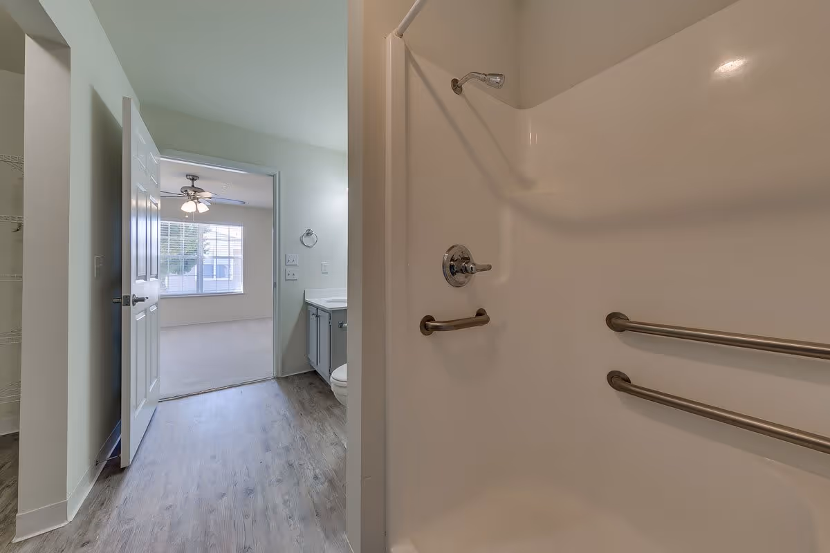 View of a bathroom with a white shower stall equipped with two horizontal grab bars and a showerhead. The bathroom has a gray vanity with a sink and a toilet visible. The door to the bathroom is open, showing a room with a ceiling fan and a large window letting in natural light.
