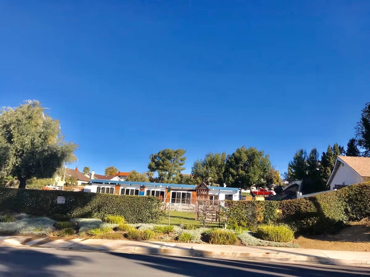 Exterior view of a single-story building with a blue roof behind a green hedge and chain-link fence, surrounded by trees and shrubs under a clear blue sky.