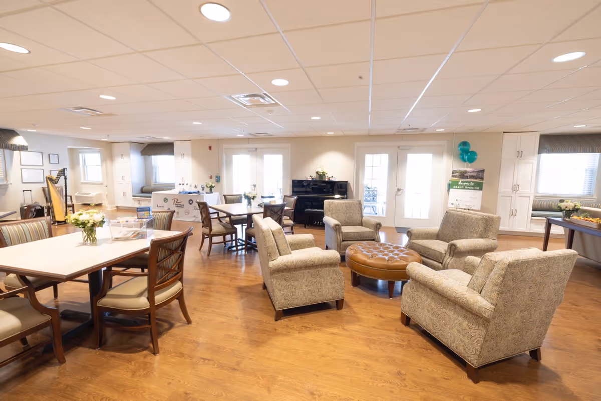 A bright and spacious common area in a retirement community featuring several upholstered armchairs arranged around a round leather ottoman, multiple tables with chairs, a piano against the far wall, and large windows and doors letting in natural light. The room has wood flooring, white walls, and ceiling lights. There are decorative flowers on the tables and a sign with balloons near the doors.