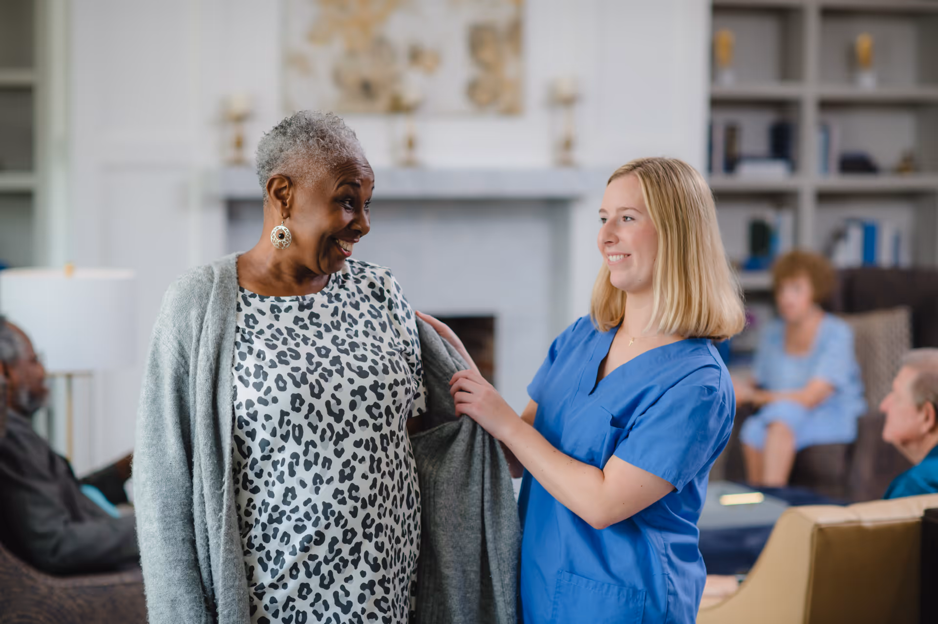 A smiling elderly woman wearing a leopard print dress is being helped with her gray cardigan by a smiling young female caregiver in blue scrubs in a cozy living room setting with other elderly people seated in the background.