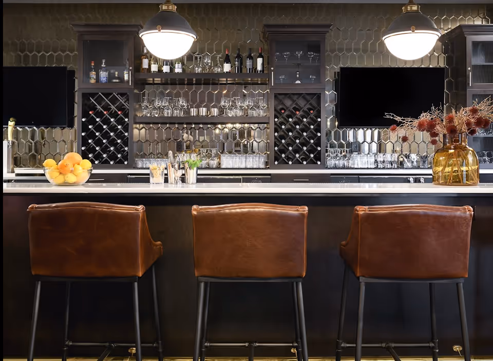 Interior bar area with three leather barstools at a marble countertop, back wall wine racks, glassware, and pendant lights.