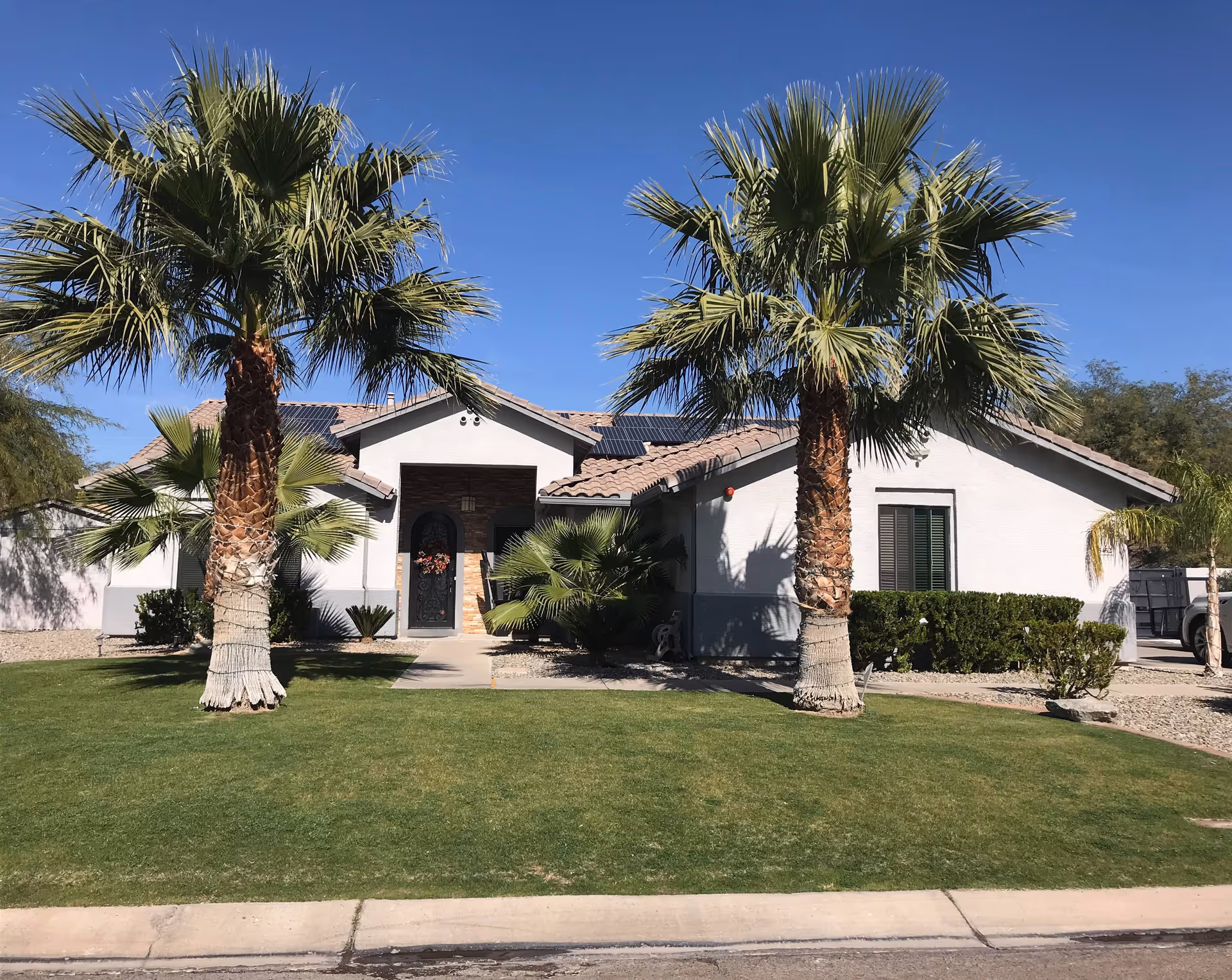 Single-story residential building with a tiled roof, white exterior walls, and a front door decorated with a wreath. The building is flanked by two tall palm trees and surrounded by green grass and shrubs under a clear blue sky.