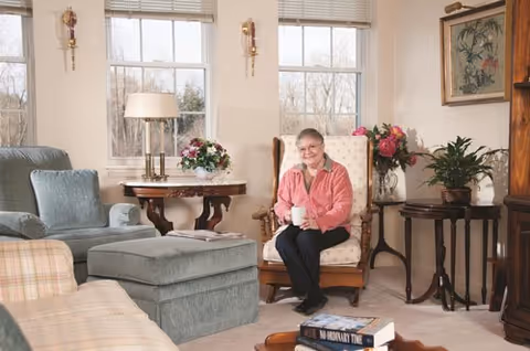 A senior woman sitting in a cushioned armchair holding a white mug in a cozy living room. The room features large windows with blinds, a side table with a lamp and a flower arrangement, another table with a potted plant and flowers, and comfortable upholstered furniture including a sofa and an ottoman. There are framed pictures on the walls and books on a coffee table in the foreground.