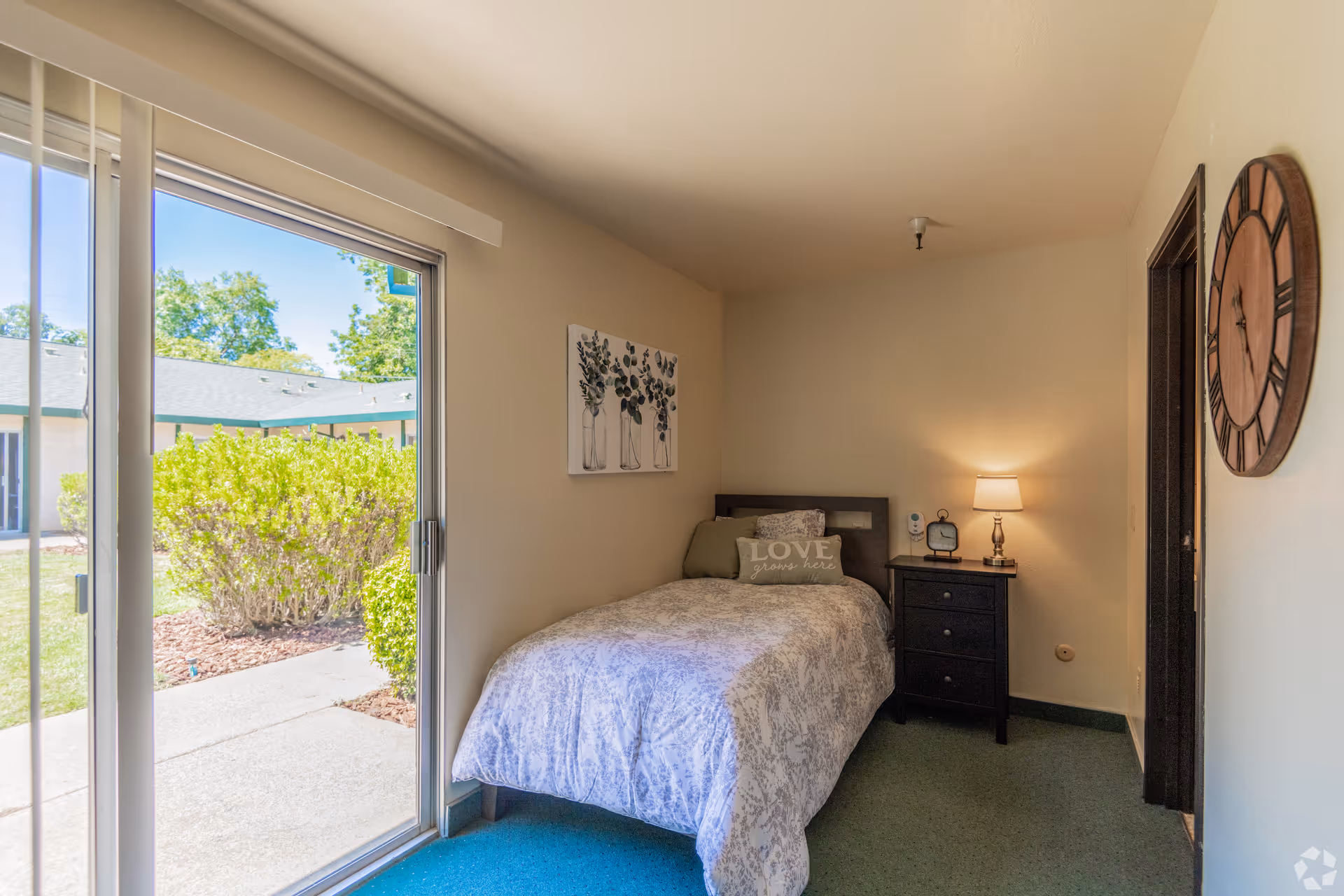 A small bedroom with a single bed covered in a patterned comforter and pillows, including one with the text 'LOVE grows here'. Next to the bed is a dark wooden nightstand with a lamp, a clock, and a small device. A large round wall clock hangs on the adjacent wall. The room has a sliding glass door that opens to an outdoor area with bushes and a building visible outside.