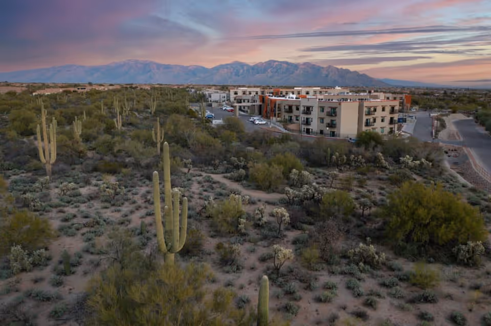 Aerial view of La Sonora at Dove Mountain senior living facility surrounded by desert landscape with cacti and shrubs, with mountains in the background under a colorful sunset sky.