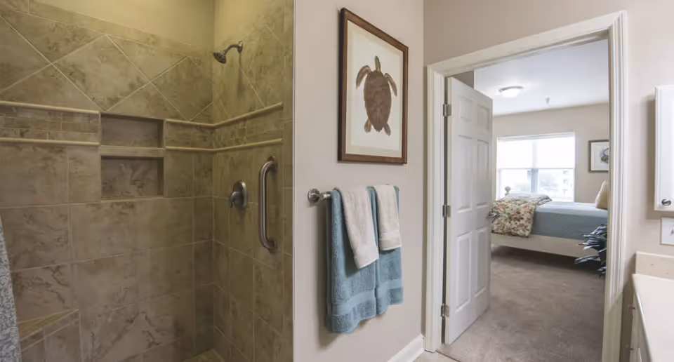 Interior view of a senior living facility bathroom with a tiled walk-in shower featuring a built-in niche and a grab bar. Two blue and white towels hang on a towel rack next to the shower. A framed turtle artwork is mounted on the wall. Through an open door, a bedroom with a bed, floral blanket, and window is visible.