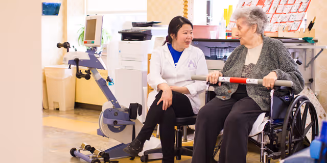 A healthcare professional in a white coat is sitting and talking with an elderly woman in a wheelchair who is holding a horizontal exercise bar in a bright room with medical equipment and office supplies in the background.