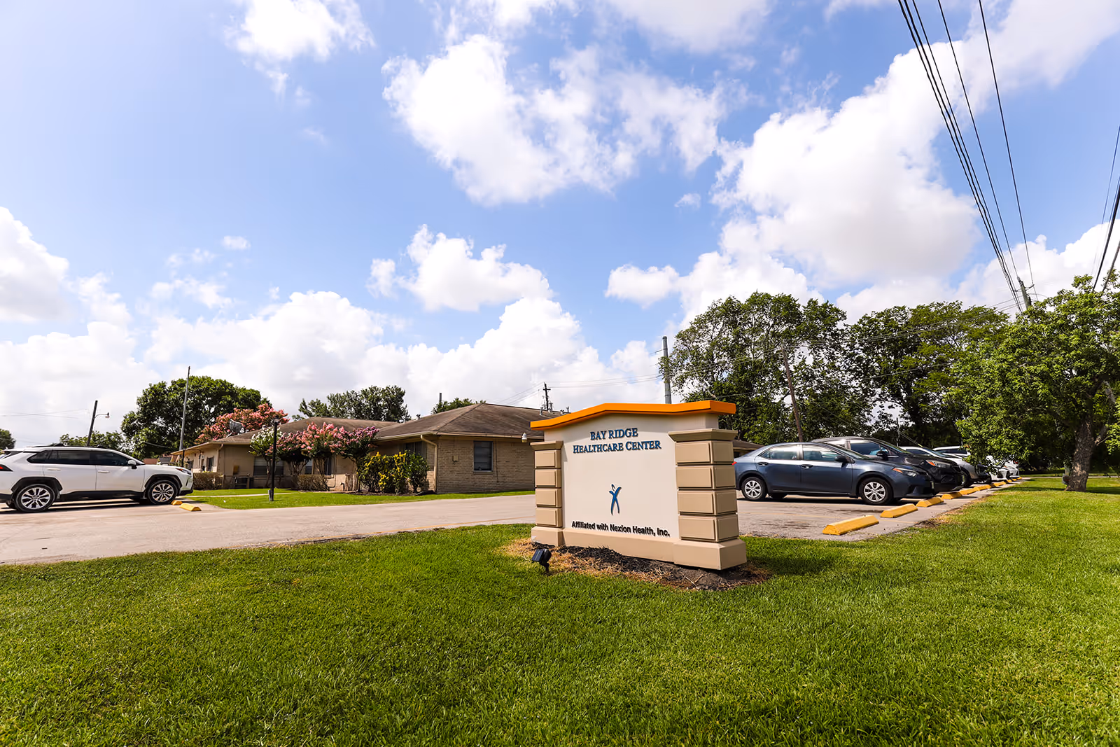 The Bay Ridge Healthcare Center sign sits on a grassy lawn in front of a single-story building and parked cars under a blue sky.