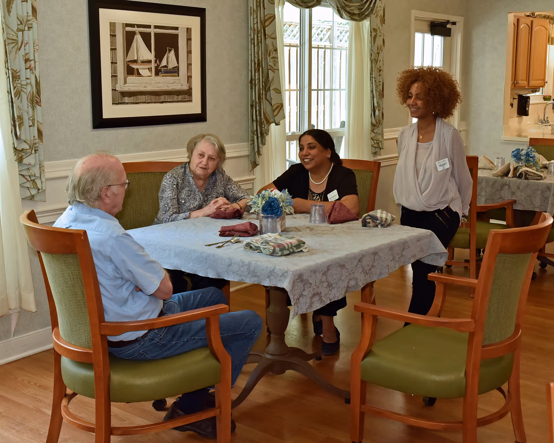 Two elderly individuals and two staff members sitting and standing around a dining table in a well-lit room with large windows and floral curtains. The table is covered with a lace tablecloth and set with napkins, utensils, and a small flower arrangement. The room has wooden floors and light-colored walls with framed artwork.