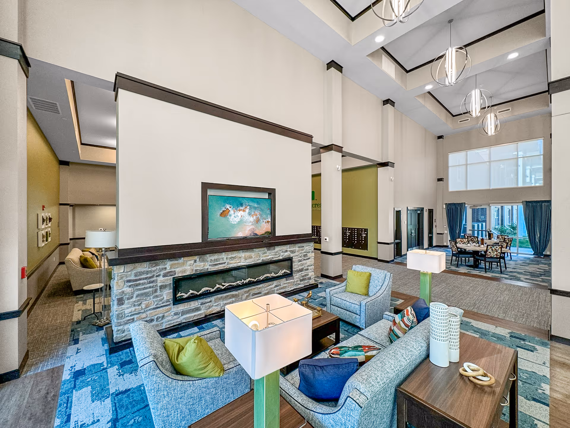 A spacious and modern senior living facility common area with high ceilings and large windows. The room features a stone fireplace with a mounted TV above it, surrounded by comfortable seating including sofas and armchairs with colorful cushions. There are decorative lamps on side tables and a dining area with a table and chairs near the windows with blue curtains. The space is well-lit with hanging spherical light fixtures and has a mix of carpet and wood flooring.