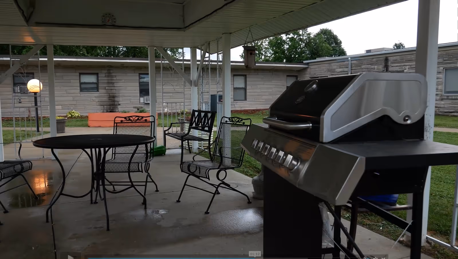Covered outdoor patio area with a round metal table, several metal chairs, and a large gas grill. In the background, there is a single-story building with windows and some greenery.