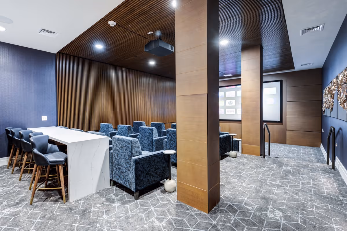 A modern media room with rows of blue upholstered armchairs facing a large screen on the wall. There is a white marble counter with black bar stools on the left side. The room features a wood-paneled ceiling and walls, with two large wooden columns in the center. The floor is carpeted with a geometric pattern.