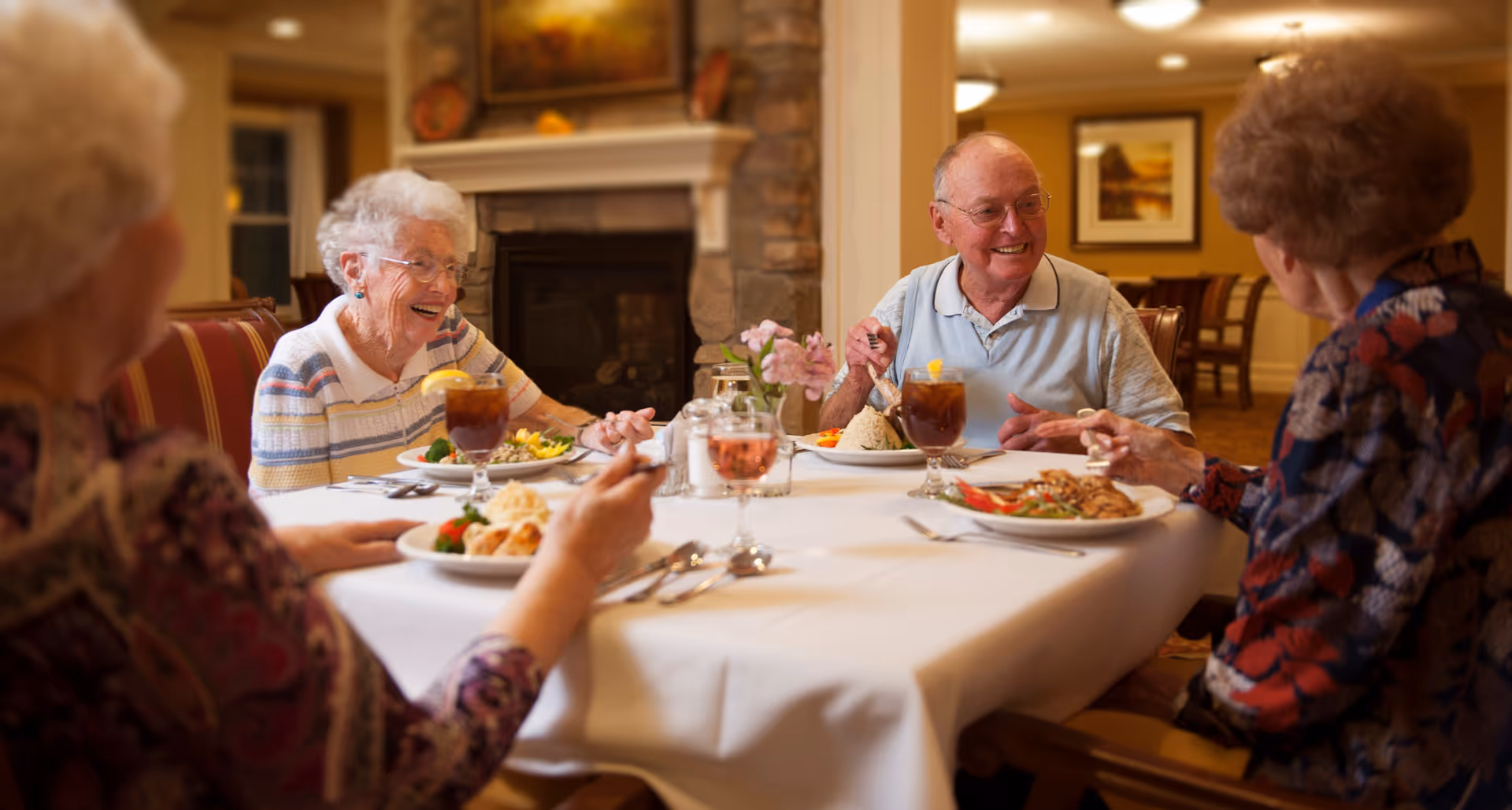 Four elderly residents sit around a table in a dining room, smiling and enjoying a meal.