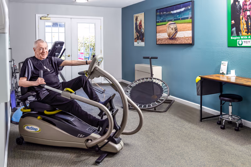 An elderly man exercising on a NuStep recumbent cross trainer in a bright fitness room with blue walls. The room features a small trampoline, a desk with a stool, and framed pictures on the wall, including a close-up of a baseball on a field. Double glass doors lead outside, allowing natural light into the space.