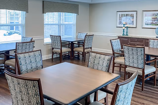 Dining room with several square wooden tables and patterned upholstered chairs near windows and a sideboard with decor.