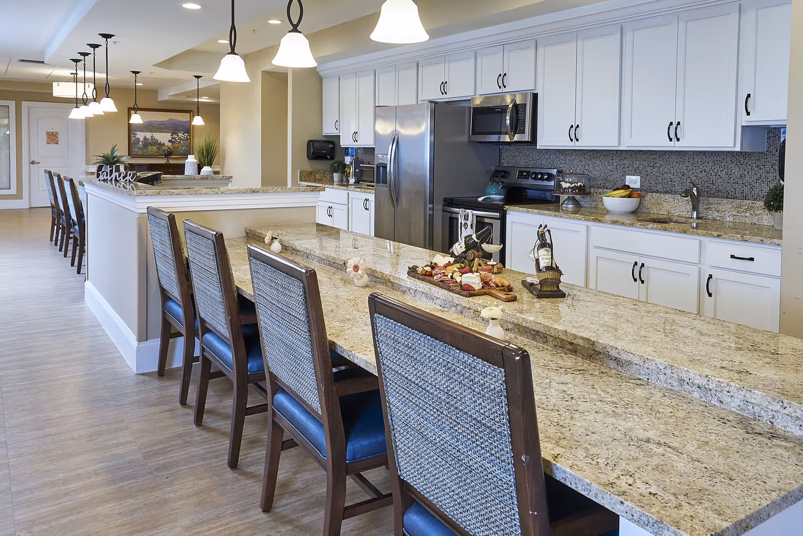 A spacious, modern kitchen area in a senior living facility with a long granite countertop island featuring several chairs. The kitchen has white cabinets, stainless steel appliances including a refrigerator, oven, and microwave. The countertop has a decorative charcuterie board and wine bottles. Pendant lights hang from the ceiling, and the floor is wood-style.