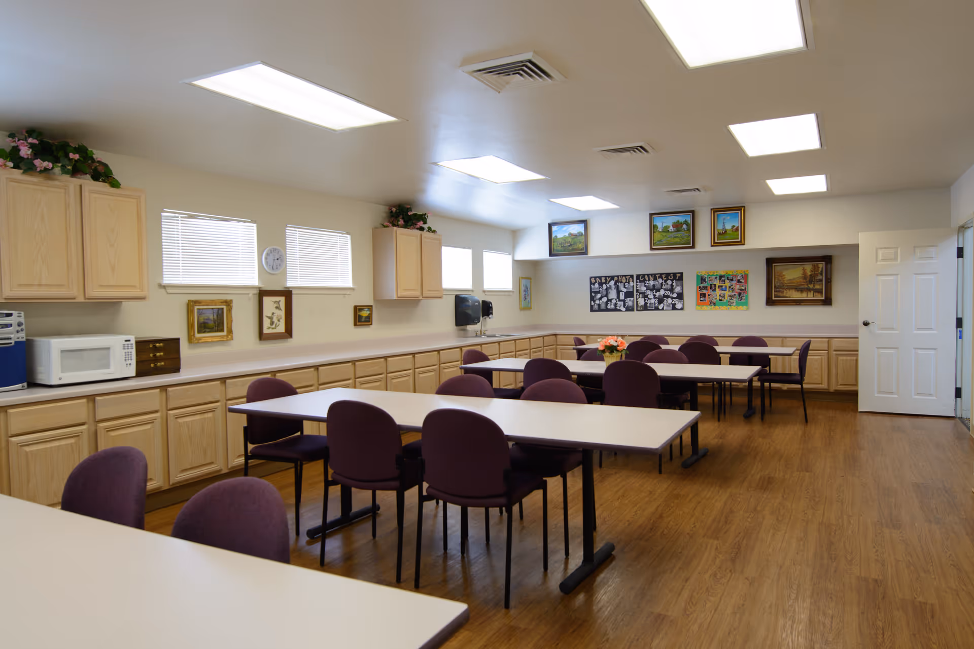 A well-lit room with multiple tables and purple chairs arranged for group seating. The room has wooden flooring, light-colored walls, and several cabinets along the walls. There are small windows near the ceiling, framed pictures on the walls, and fluorescent ceiling lights. A microwave and a small stereo are placed on the countertop, and a flower arrangement is on one of the tables.