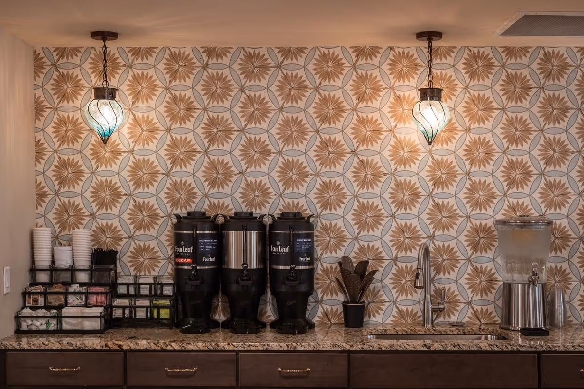 A beverage station with three large coffee dispensers labeled Four Leaf, a water dispenser, a sink with a faucet, and various coffee supplies including cups, stirrers, and sweeteners on a granite countertop. The wall behind features a decorative tile pattern with brown and beige floral designs, and two hanging pendant lights with green and white glass shades.
