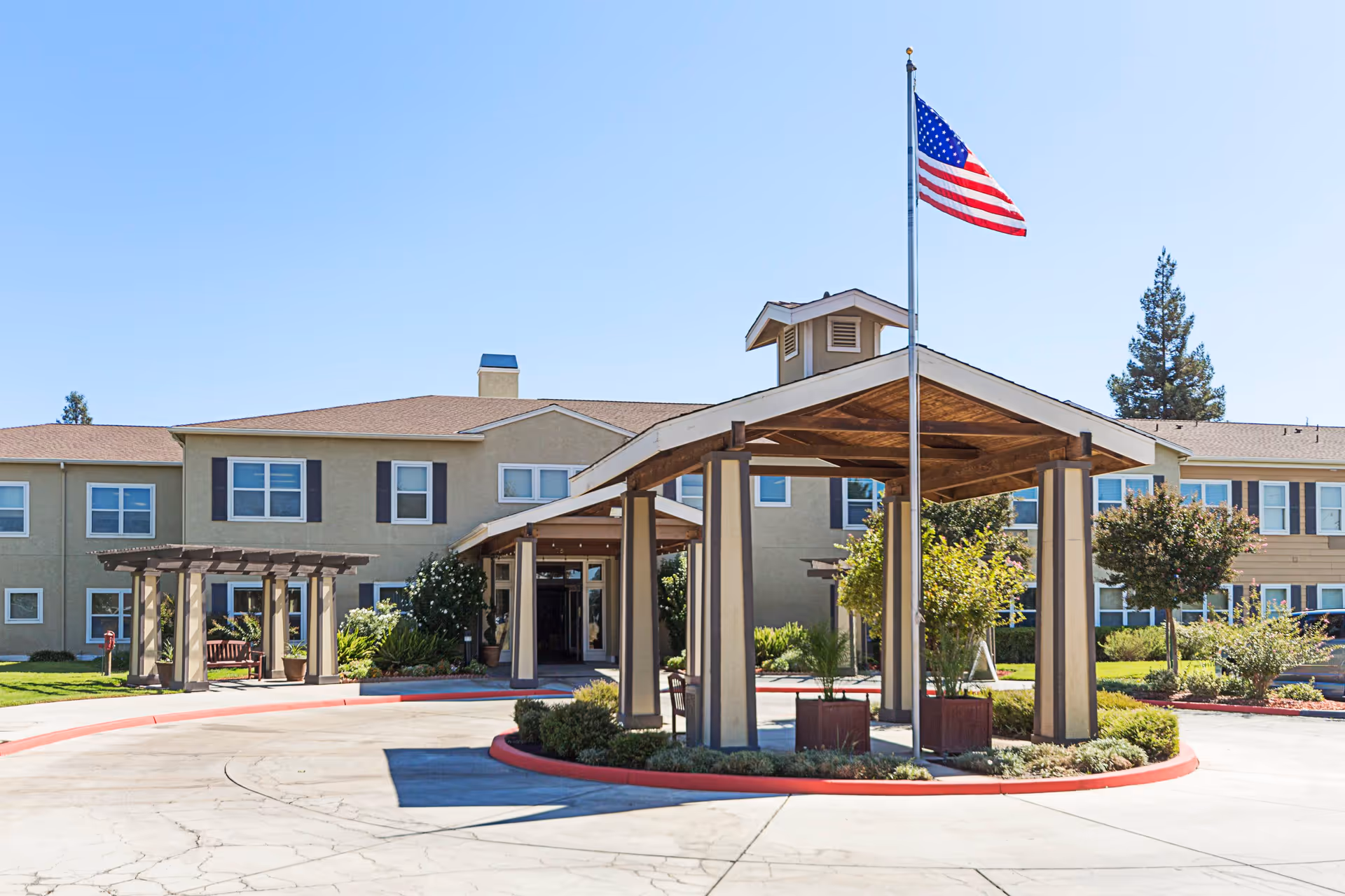 Exterior view of Truewood by Merrill, Clovis senior living facility showing a two-story building with multiple windows, a covered driveway entrance with wooden beams, an American flag on a flagpole, and landscaped greenery under a clear blue sky.