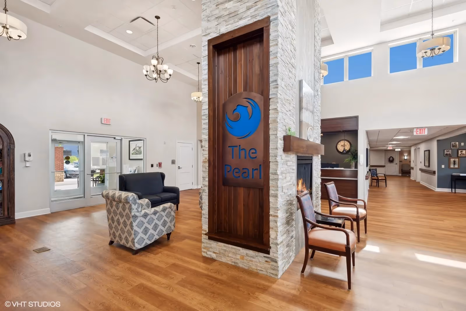 Bright, open senior living lobby with chairs and couches around a central stone fireplace featuring a wooden sign reading "The Pearl".