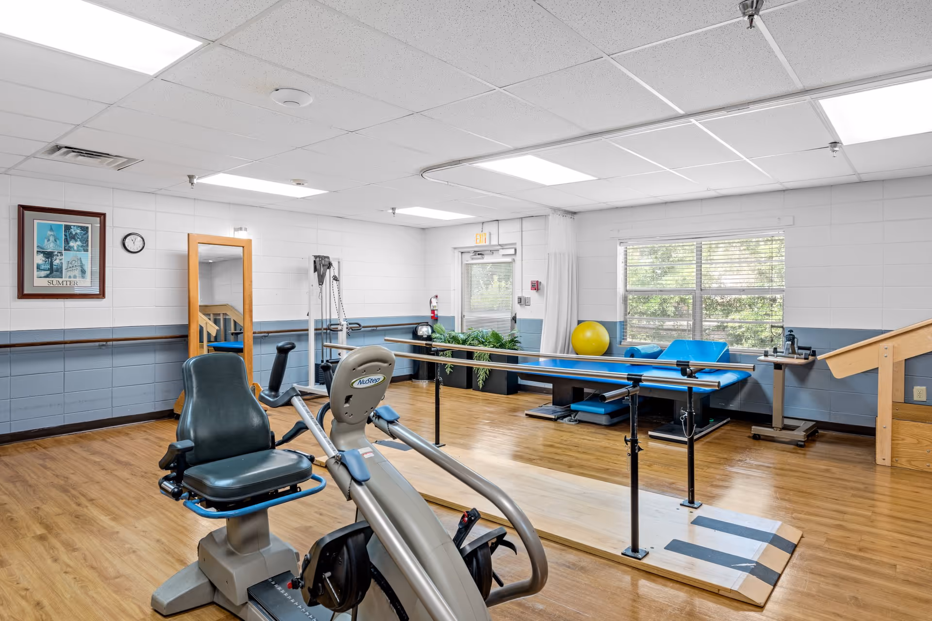 A rehabilitation room with exercise equipment including a stationary recumbent bike, parallel bars for walking practice, a blue therapy table, a yellow exercise ball, and a wooden staircase for physical therapy. The room has wood flooring, white and blue walls, a large window, and fluorescent ceiling lights.