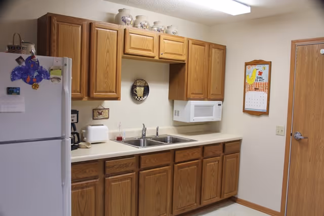 A kitchen area with wooden cabinets above and below a countertop. The countertop has a double sink, a white microwave mounted on the wall, a toaster, a coffee maker, and a soap dispenser. A white refrigerator with magnets and papers is on the left side. A calendar with a chicken design is hanging on the wall next to a wooden door.