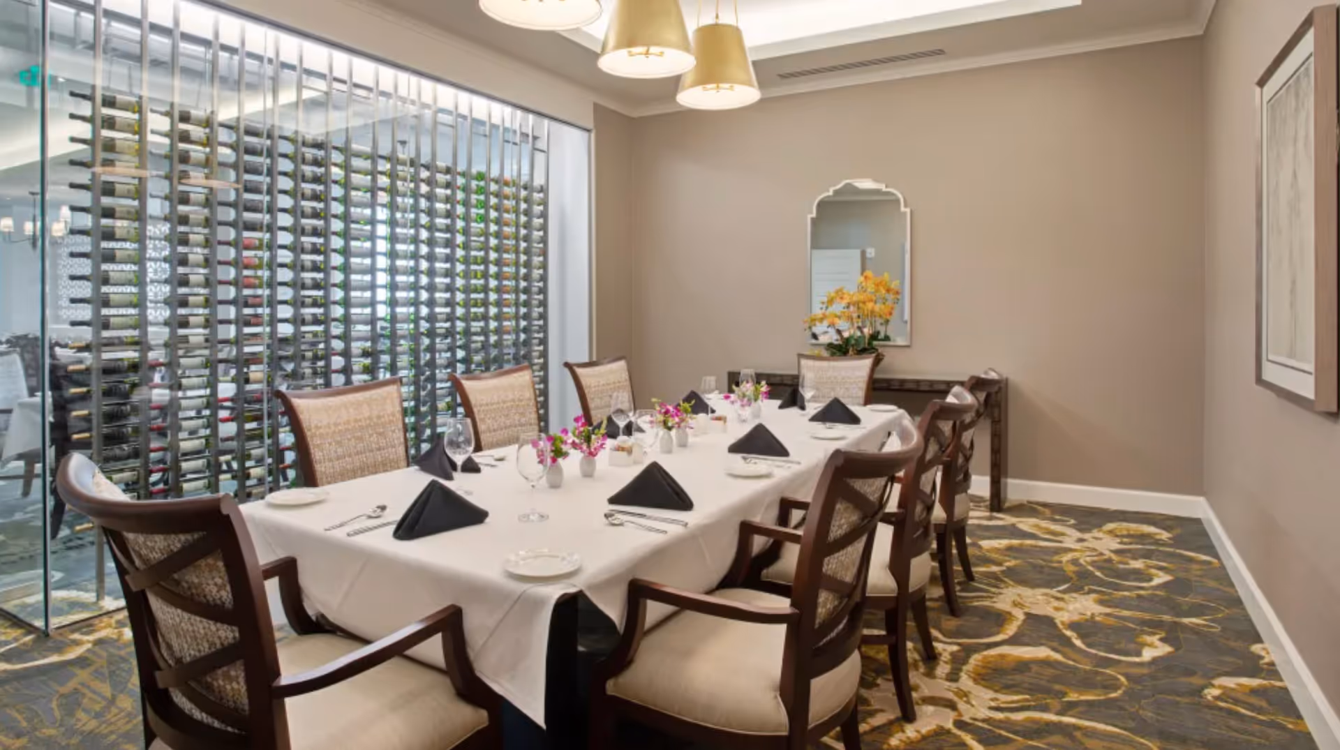 A formal dining room with a long rectangular table covered with a white tablecloth, set with black folded napkins, glassware, and small floral centerpieces. The room features wooden chairs with cushioned seats and backs, a decorative mirror on the wall, and a glass wall displaying a large wine rack filled with bottles. The carpet has a patterned design in shades of gray and yellow, and three pendant lights hang from the ceiling.