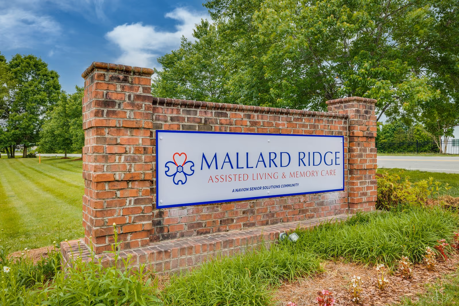 A brick sign for Mallard Ridge Assisted Living & Memory Care, a Navion Senior Solutions community, set in a landscaped area with green grass, plants, and trees under a partly cloudy blue sky.