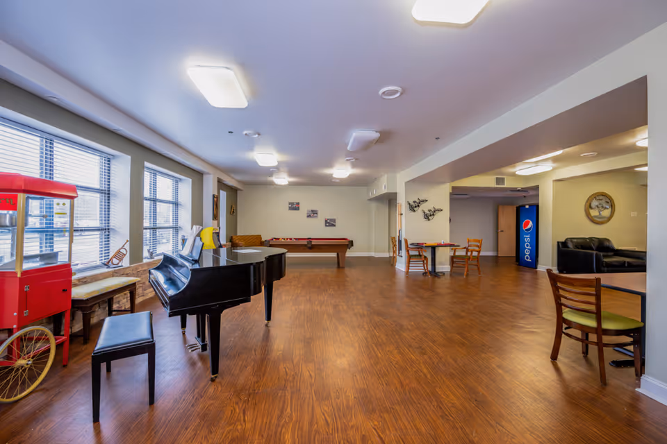 A spacious common area in a senior living facility featuring a black grand piano with a bench, a red popcorn machine, a pool table, several wooden chairs and tables, a Pepsi vending machine, and a black leather couch. The room has large windows with blinds, wood flooring, and bright ceiling lights.