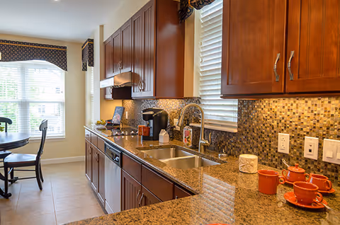 Sunlit kitchen with granite countertops, dark wood cabinets, a mosaic tile backsplash, double sink, coffee maker, and a small dining table by a window.