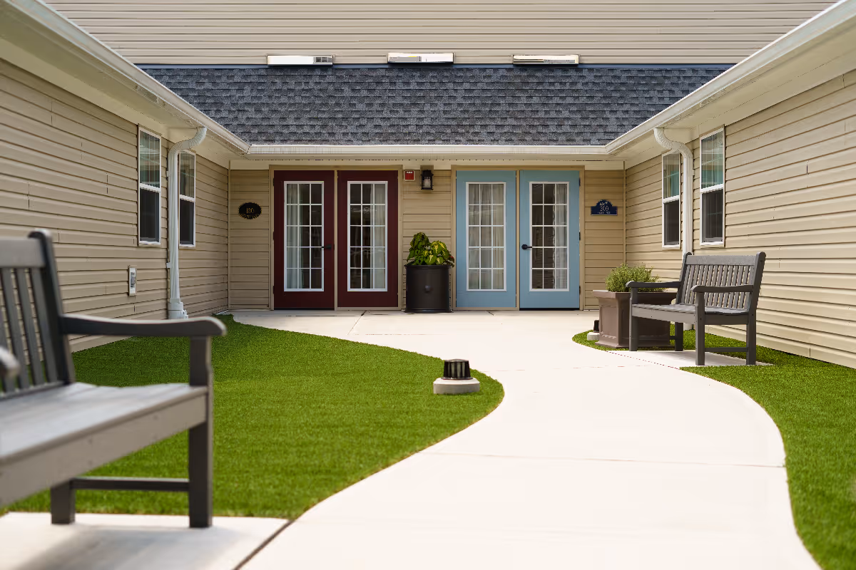 A courtyard area with a curved concrete walkway surrounded by green artificial grass. There are two benches on either side of the walkway and potted plants near the building. The building has beige siding with two sets of French doors, one set painted red and the other blue.