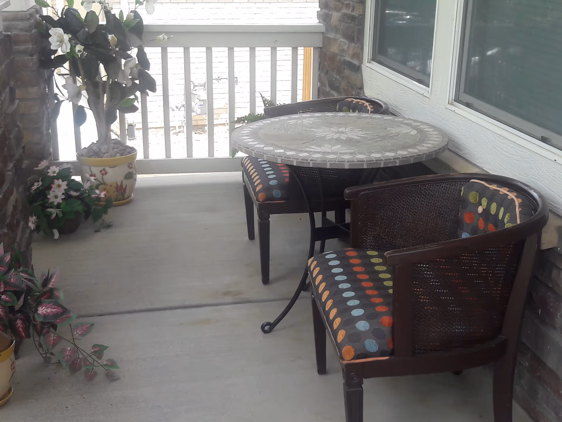 A small outdoor patio area with a round mosaic table and two chairs with colorful polka dot cushions. There are several potted plants with flowers placed along the edges of the patio. The patio is enclosed with a white railing and has a brick wall on one side and a window on the other.