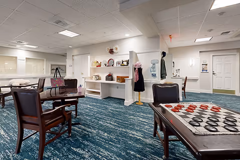 A spacious common area with blue patterned carpet, several wooden tables and chairs, and a checkers game set up on one table. The walls are white with built-in shelves displaying decorative items. There are doors and a mannequin with a hat and scarf near the center of the room. The ceiling has recessed lighting and air vents.